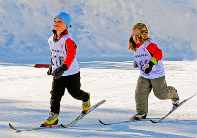 Two youth skiing in a line without poles wearing jerseys.